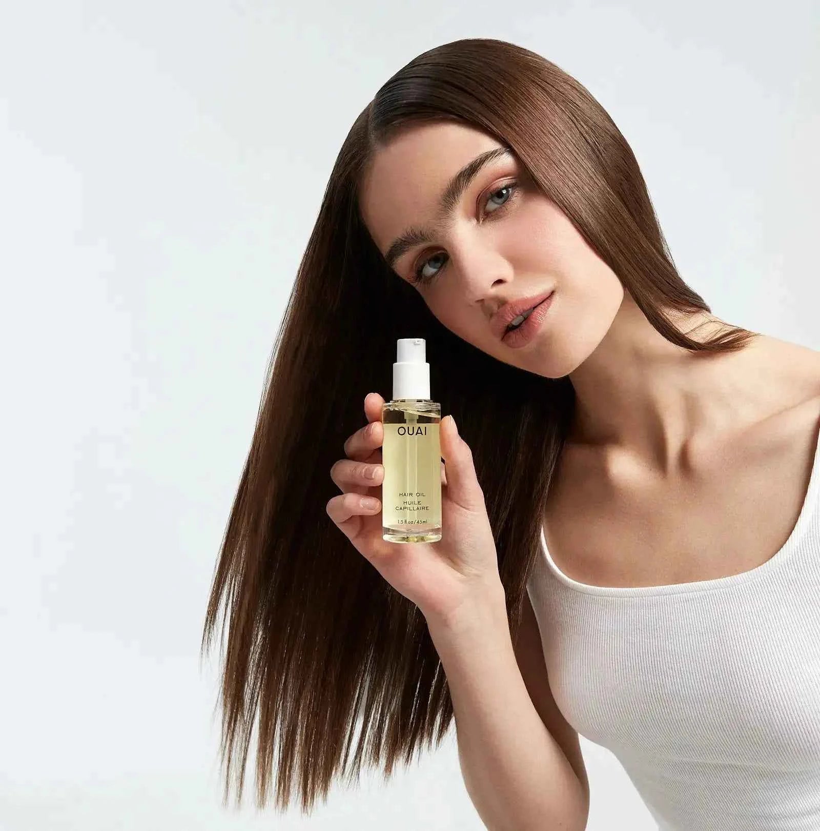 Ouai hair oil bottle held near long brown hair, pump-top visible